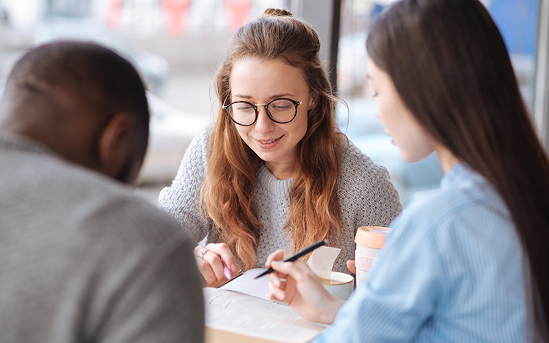 Three people sit at a desk, looking at some kind of material. One of them sits with their face towards the camera and is in focus. The other two people sit with their backs towards the camera and appear blurry.